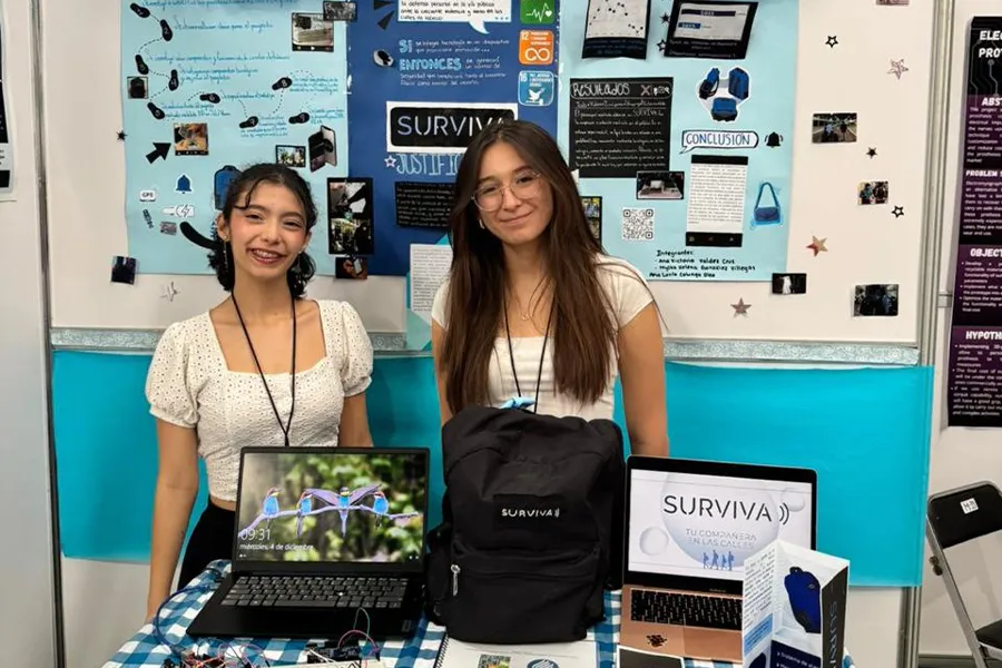 Dos alumnas de preparatoria posan en stand de feria de ciencias mostrando una mochila.