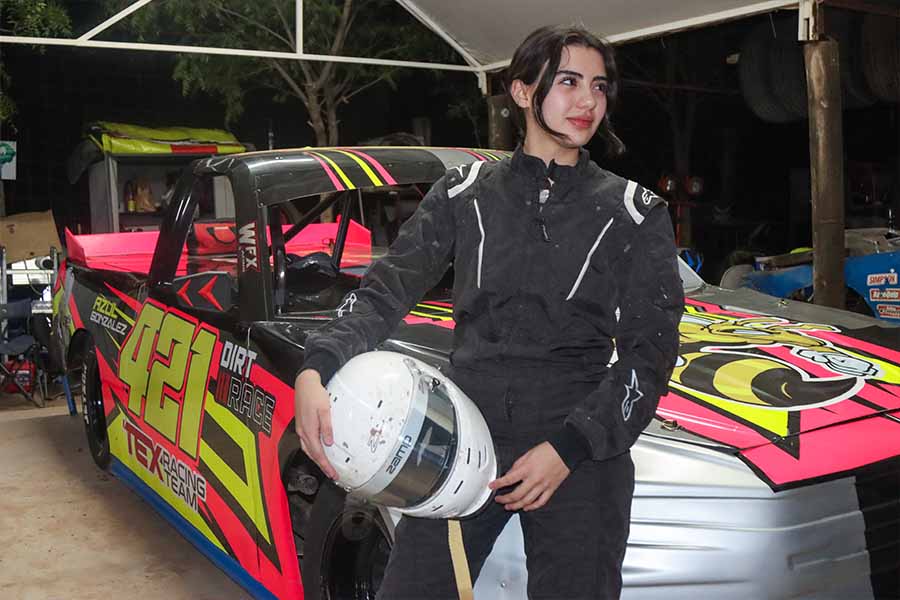 Mujer joven posa en uniforme de piloto automovilístico junto a su carro.