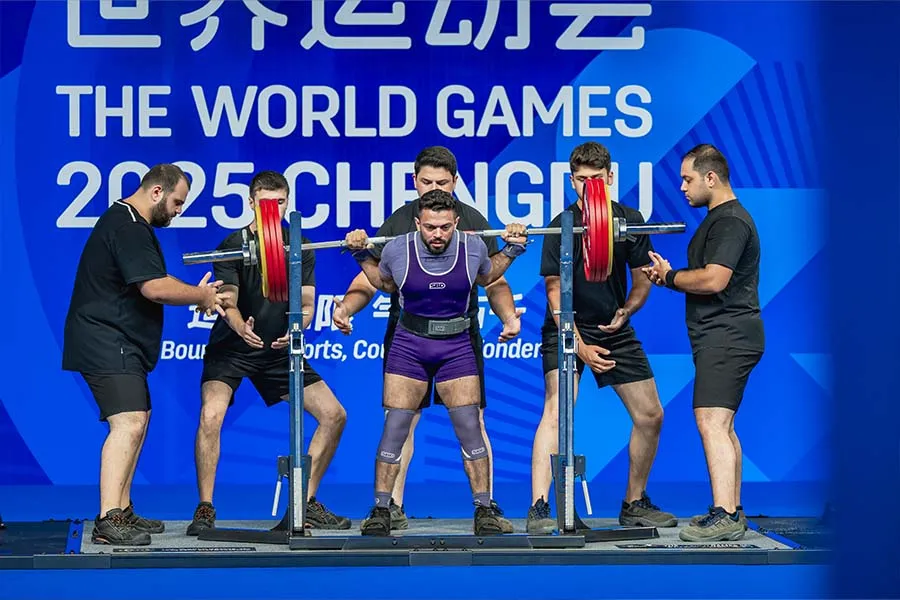 Hombre sosteniendo pesas sobre los hombres rodeado de spotters en el escenario de una competencia mundial.