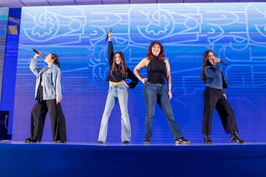 4 jóvenes sobre un escenario con pantallas de fondo sosteniendo micrófonos.