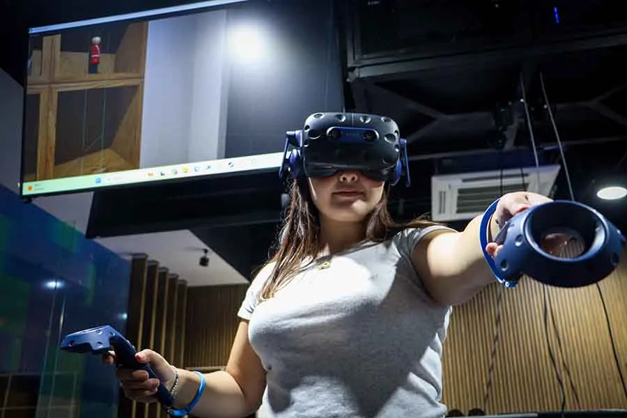 Una chica portando con visor de realidad virtual sostiene en sus manos dos controles. En el fondo cuelga una pantalla del techo que muestra la simulación que ella experiemnta.
