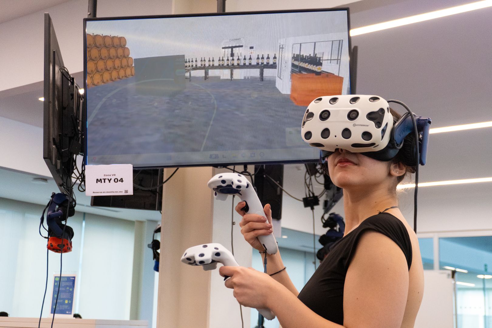 Estudiante del Tec usando visor de realidad virtual y controladores mientras interact&uacute;a con una simulaci&oacute;n digital de una planta tequilera mostrada en una pantalla.