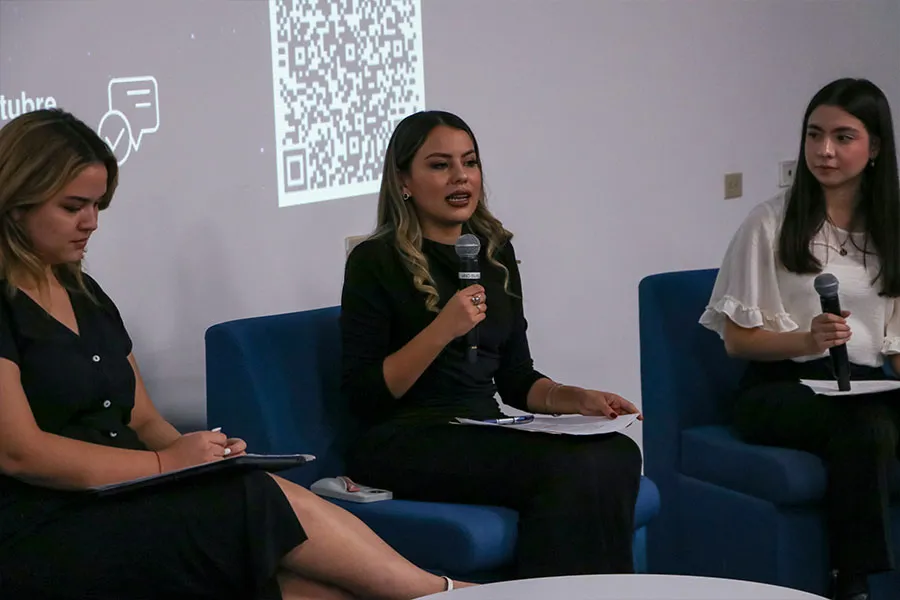 Tres mujeres sentadas en sillones azules mientras la persona del centro habla sosteniendo un micrófono.