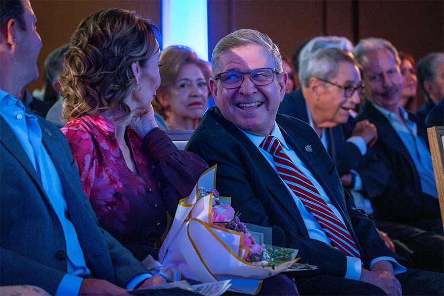 Público sentado en sillas. Al centro está enfocando un hombre riéndose y volteando hacia la mujer que está sentada a su lado. 