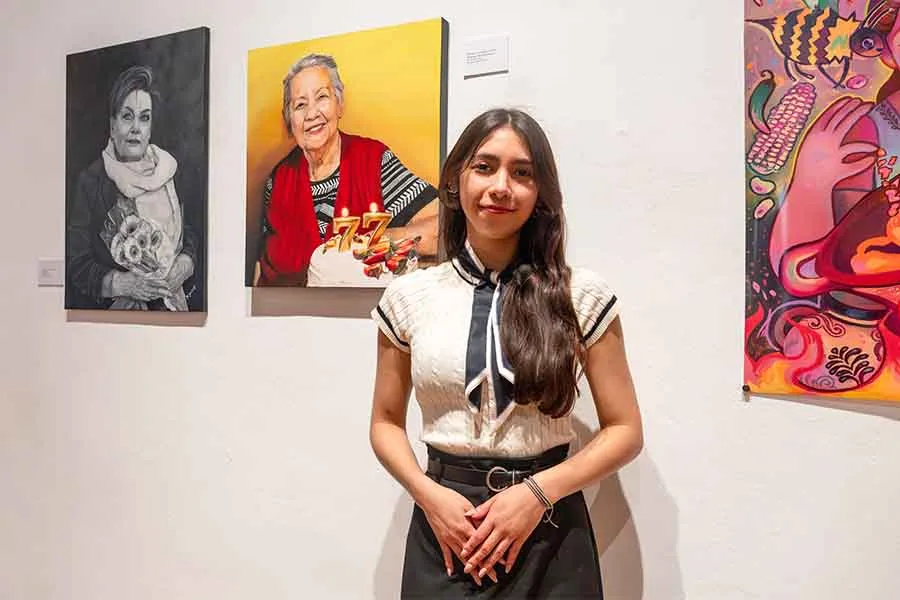 Mujer adolescente posando frente a una pintura que muestra a una mujer mayor con un pastel que tiene unas velas en forma del número 77.