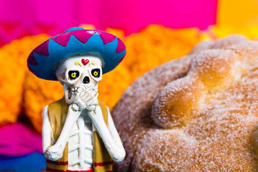 Decoración de calavera tapándose la boca a un lado de un pan de muerto.