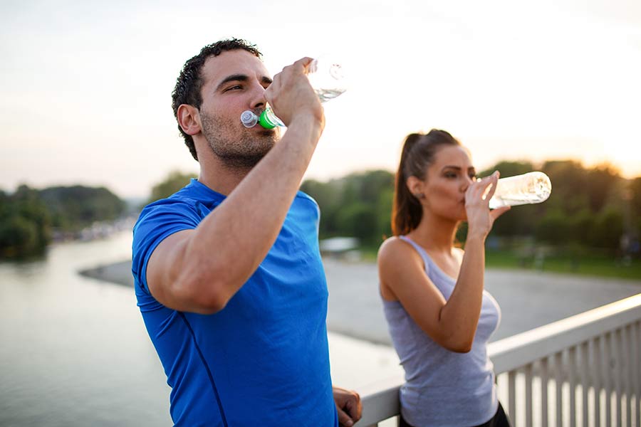 Hombre y mujer durante una pausa en su rutina de ejercicios al aire libre; se observan hidratándose y con ropa clara, recomendaciones para evitar un golpe de calor