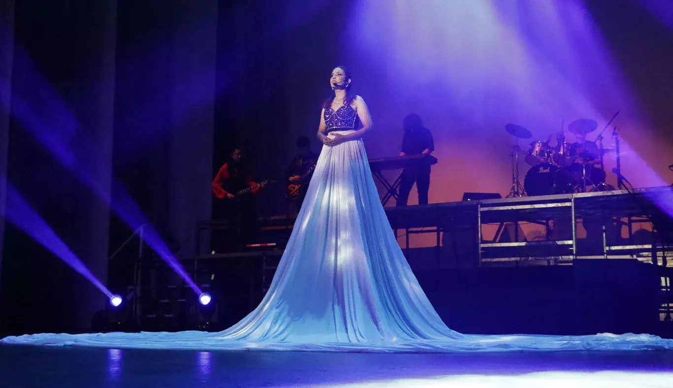 Chica cantante en el centro del escenario con una falda larga blanca sobre la cual se proyectan estrellas.  