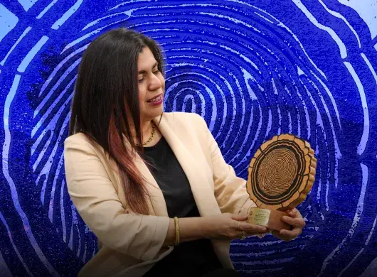 Mujer con saco blanco sosteniendo trofeo con forma de corte transversal de tronco y mirando hacia el.