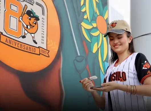 Chica portando una jersey y gorra del equipo de béisbol de Naranjeros de Hermosillo, sosteniendo una brocha con pintura blanca enfrente de un mural que muestra una pelota naranja con el logo del 80 aniversario del equipo. 