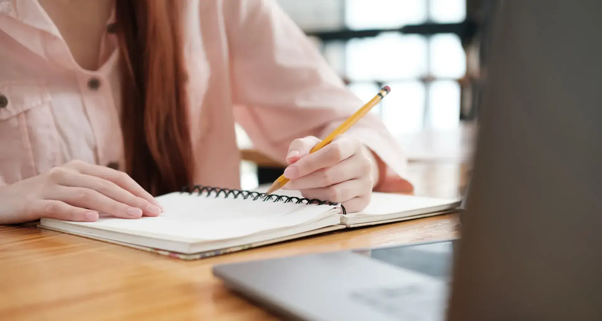 Mujer escribe en su libreta mientras observa su computadora portátil.