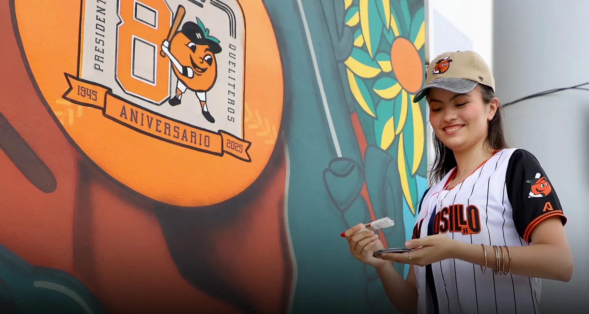 Chica portando una jersey y gorra del equipo de béisbol de Naranjeros de Hermosillo, sosteniendo una brocha con pintura blanca enfrente de un mural que muestra una pelota naranja con el logo del 80 aniversario del equipo. 