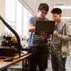 2 jóvenes estudiantes en un laboratorio; uno de ellos sostiene una laptop en su mano y señala su pantalla