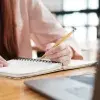 Mujer escribe en su libreta mientras observa su computadora portátil.