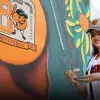 Chica portando una jersey y gorra del equipo de béisbol de Naranjeros de Hermosillo, sosteniendo una brocha con pintura blanca enfrente de un mural que muestra una pelota naranja con el logo del 80 aniversario del equipo. 