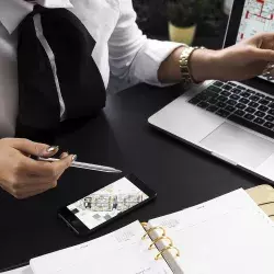 mujer con camisa blanca trabajando con computadora, celular y libretas.