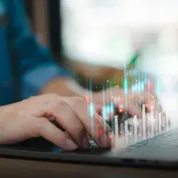 Manos sobre el teclado de una computadora con un holograma de gráficas por enfrente. 