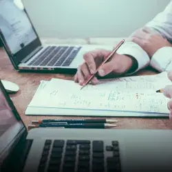 Toma cerrada de las manos de dos personas escribiendo sobre un papel con laptops a los lados. 