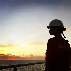 Ashly Hinojos con casco y uniforme naranja mirando el atardecer sobre el mar desde una plataforma petrolera.