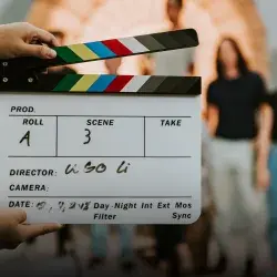 Primer plano de una claqueta de cine, al fondo se ve un grupo de personas desenfocadas.