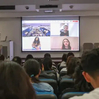 Público en auditorio volteando hacia una pantalla que muestra una videollamada en proceso.