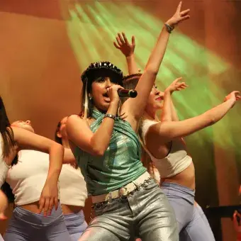 Chica con blusa brillante verde y boina canta rodeada de bailarinas vestidas de blanco.