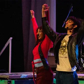 Un chico y una chica en escenario se toman de la mano con brazos alsados para saludar al público.