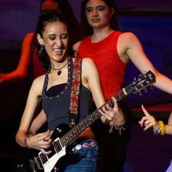 Chica tocando la guitarra eléctrica en escenario con humo alrededor. 
