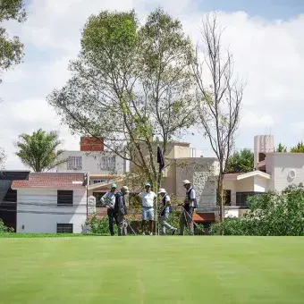 Grupo de golfistas reunidos en el green junto al banderín, con casas residenciales al fondo y árboles alrededor, durante el torneo EXATEC Blue Open en el Club de Golf Pachuca en apoyo a Líderes del Mañana.