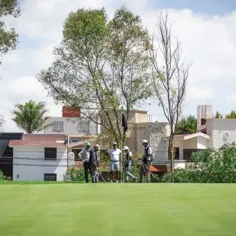 Grupo de golfistas reunidos en el green junto al banderín, con casas residenciales al fondo y árboles alrededor, durante el torneo EXATEC Blue Open en el Club de Golf Pachuca en apoyo a Líderes del Mañana.