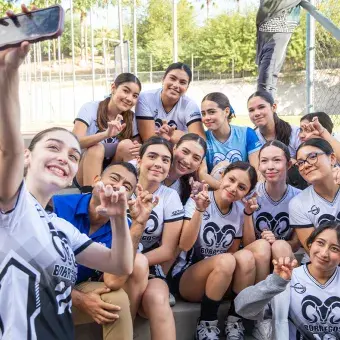 Chicas de un equipo femenil de voleibol sentadas en unas escaleras con su entrenador mientras una sostiene su celular a lo alto para tomar una foto del grupo en modo selfie. 