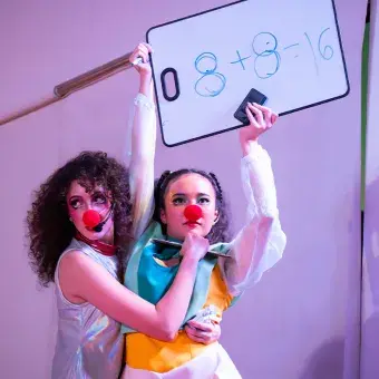 Dos actrices con vestuarios y narices de payaso, una sostiene en lo alto un letrero mientras que la otra la rodea con los brazos. 