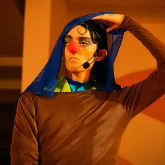 Actor con vestuario y nariz de payaso levantando un velo que le cubre la cabeza.