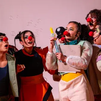 Actores con vestuarios y narices de payaso alrededor de una chica que sopla burbujas de jabón.