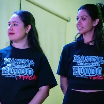 Dos jóvenes portando camisetas negras con el título de la obra Manual Para No Hacer Ruino (y Fallar).