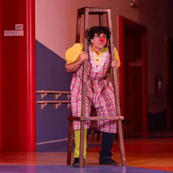 Actor con vestuario y nariz de payaso asomándose entre una escalera.